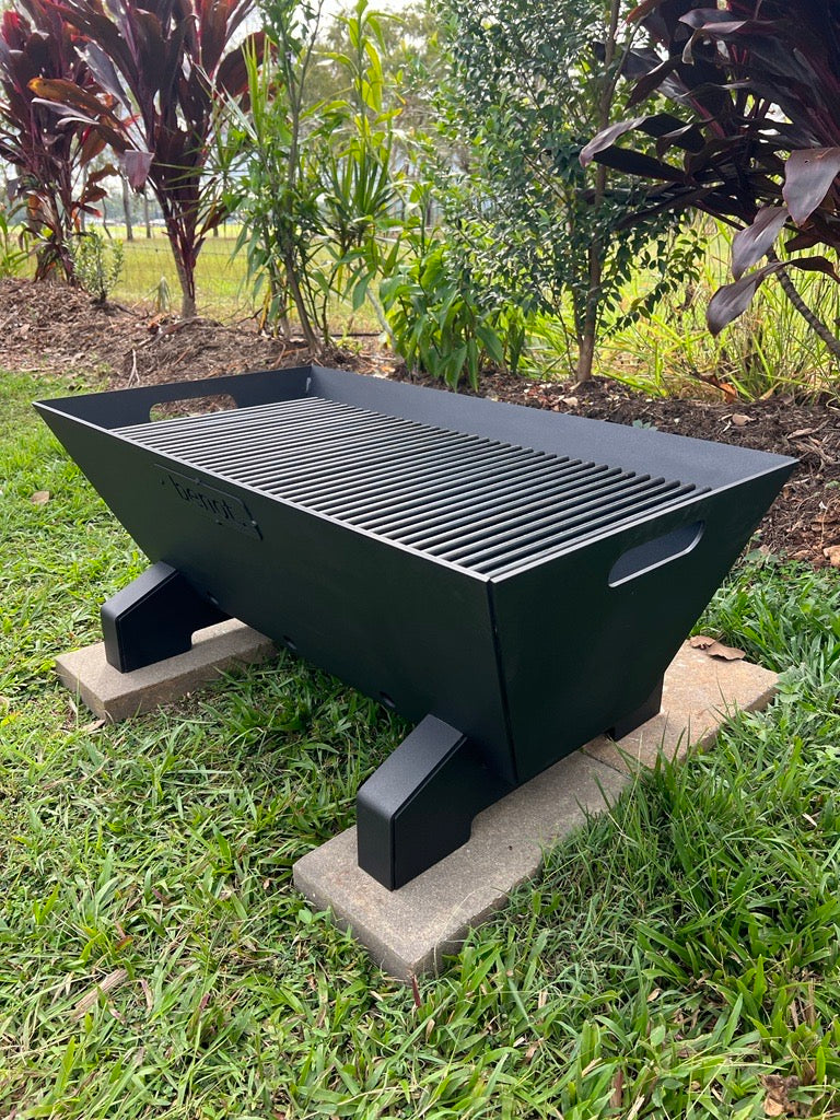 Black outdoor Fire pit bbq grill on a grassy area with plants in the background